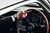 Close-up of a hand on a wood-rimmed steering wheel inside an Aston Martin, showing the dashboard and gauges.